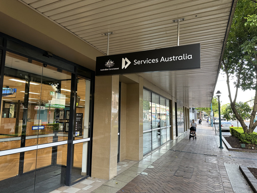 The entrance of a Services Australia service centre with glass doors and a black overhead sign on a shopping street
