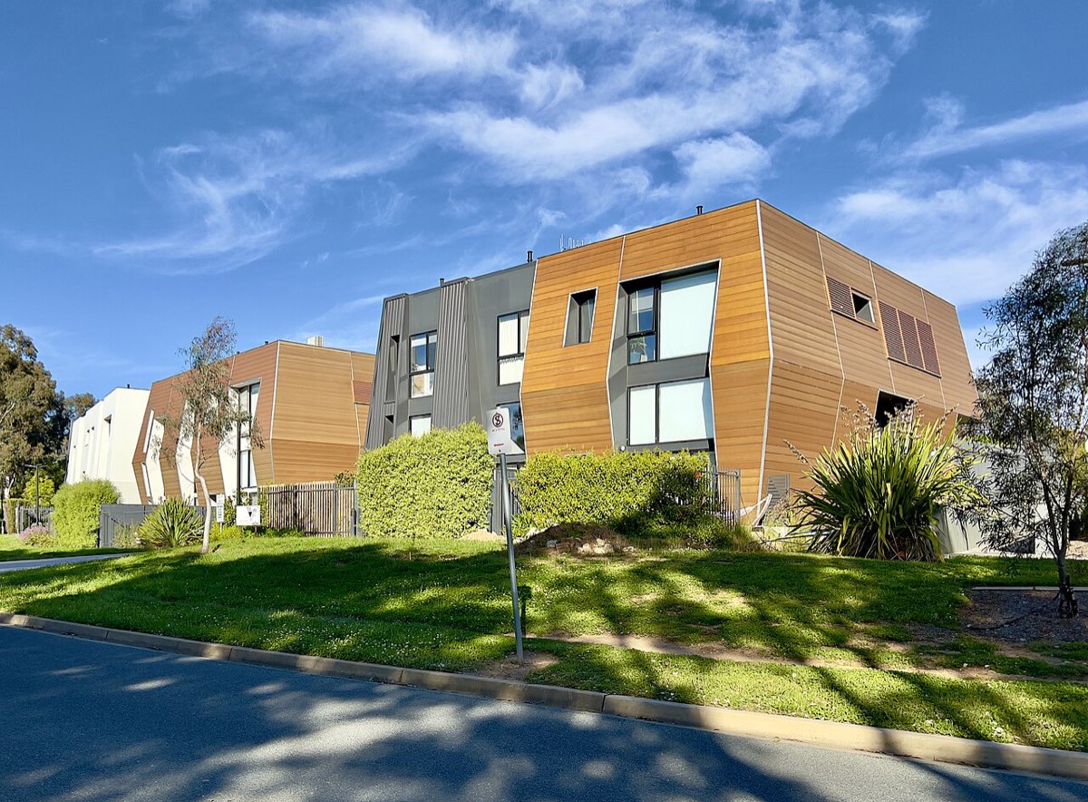 A modern two-storey office building clad in timber-coloured panels with angled walls, behind a lawn and shrubs on a sunny day