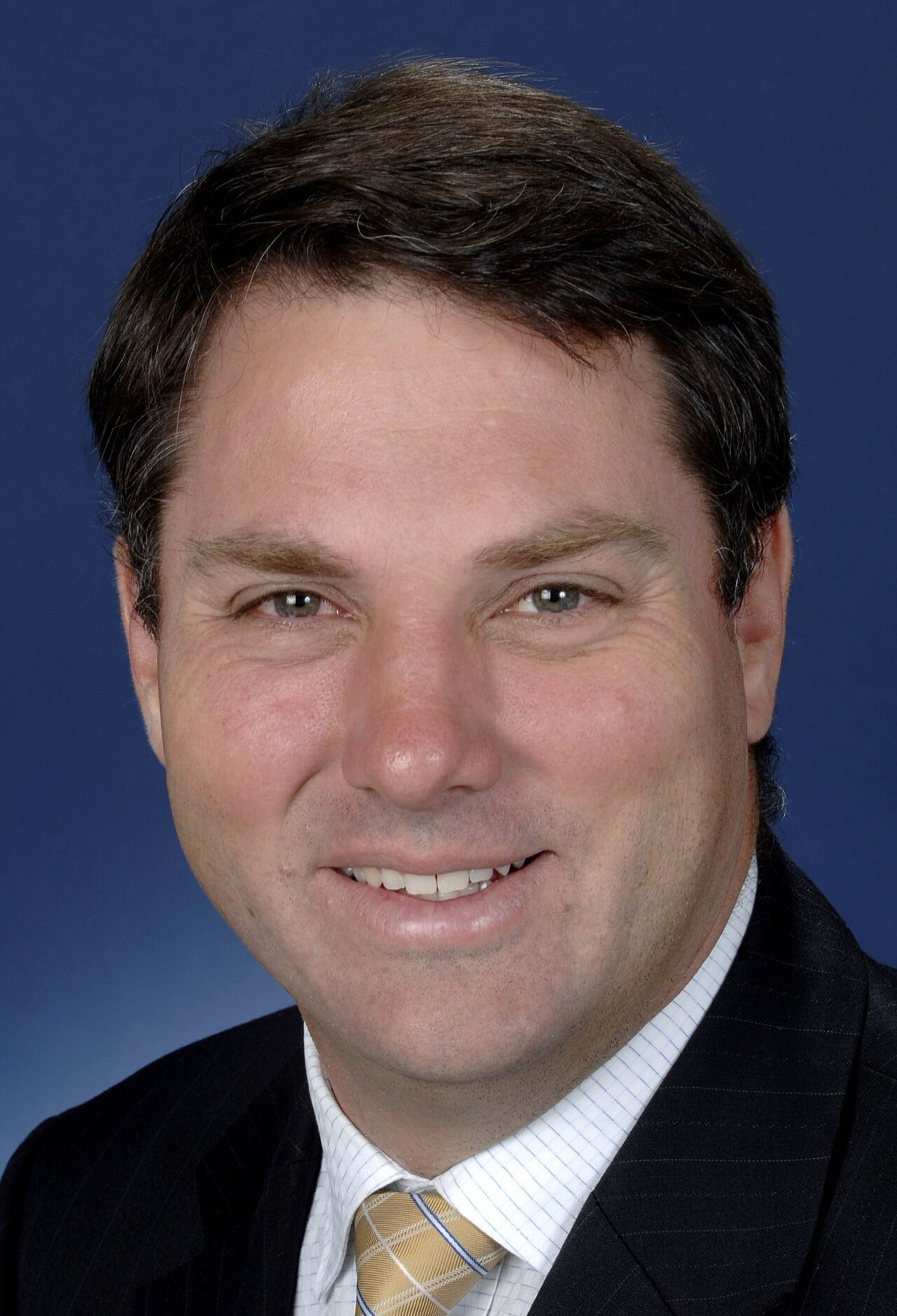 Portrait of Richard Marles, a smiling man with short dark hair in a dark suit and gold-striped tie, against a blue background