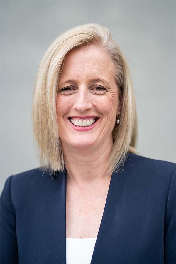 Head-and-shoulders portrait of Katy Gallagher, a smiling woman with shoulder-length blonde hair in a navy jacket, against a grey background