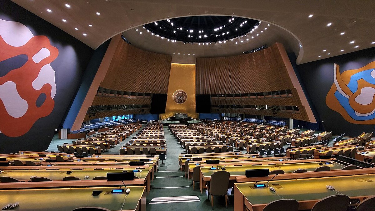 The United Nations General Assembly hall in New York, with tiers of delegate desks facing a podium under a large gold emblem
