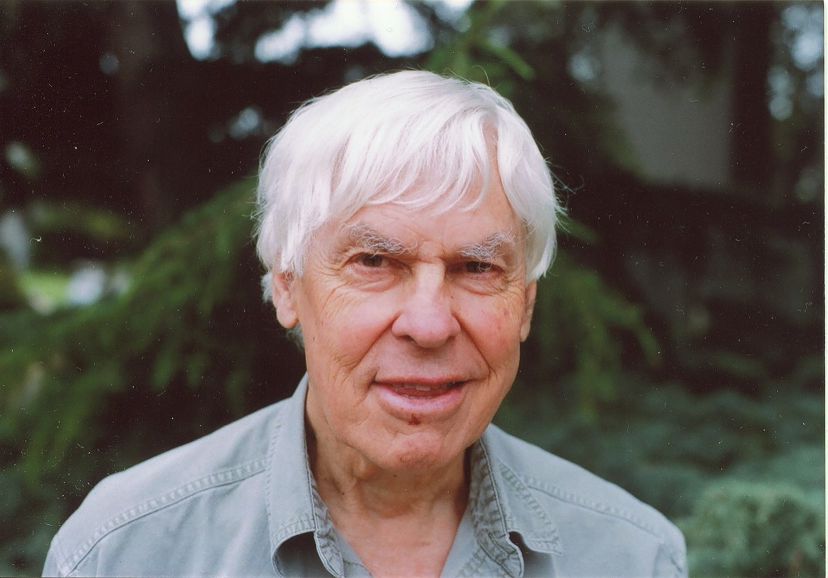 Portrait of an older man with white hair in a gray shirt, standing in front of trees