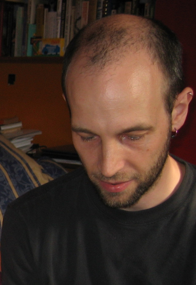 A man with close-cropped hair, a short beard and a dark t-shirt looking down, with bookshelves behind him