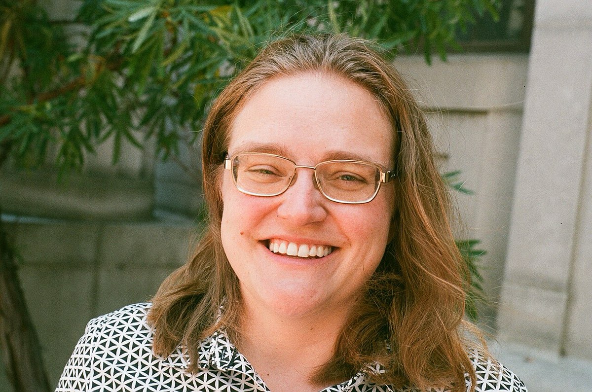 Melanie Matchett Wood, smiling, with shoulder-length hair and glasses, outdoors in front of a stone wall and greenery