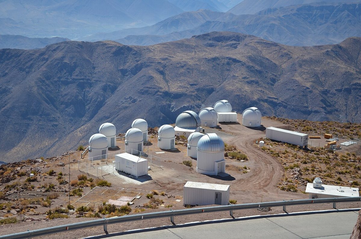 Several white observatory domes on a dry, rocky mountaintop with brown ridges and valleys behind them