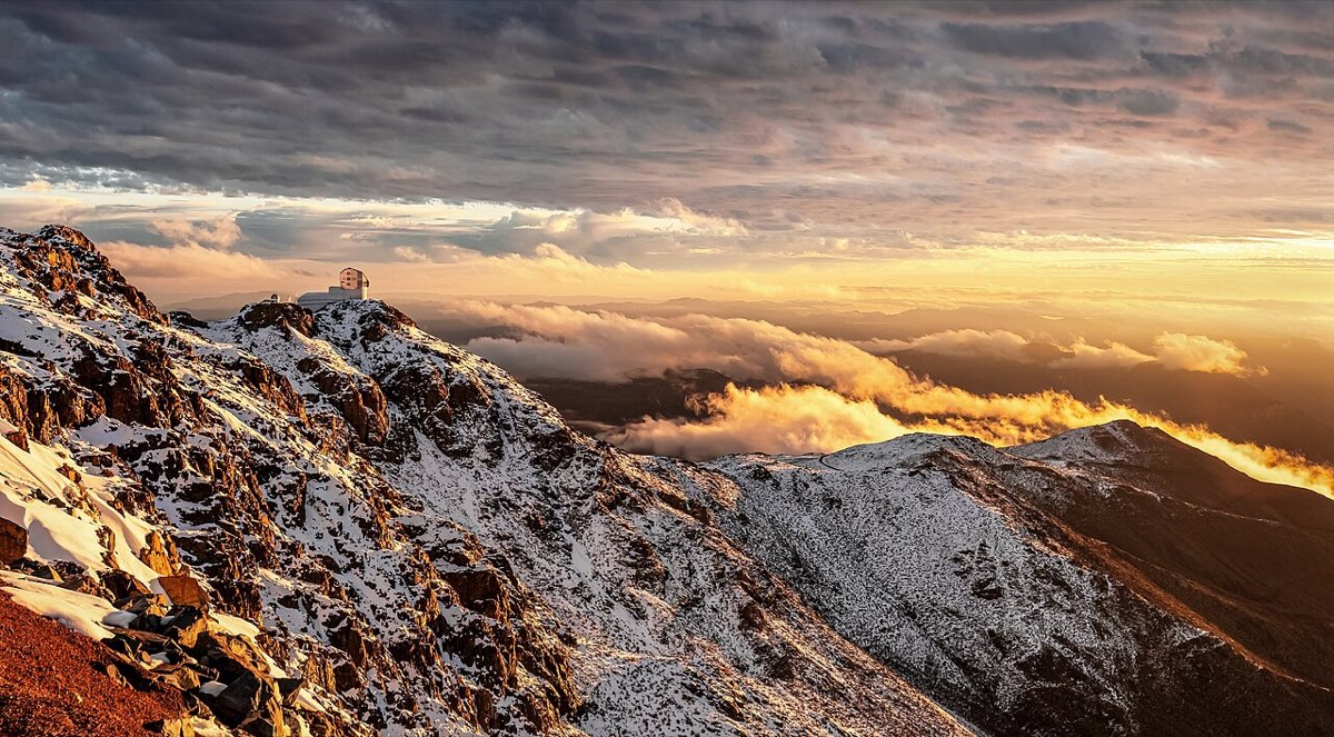 The Rubin Observatory building on a snowy mountain ridge under a dramatic orange sunset sky above the clouds
