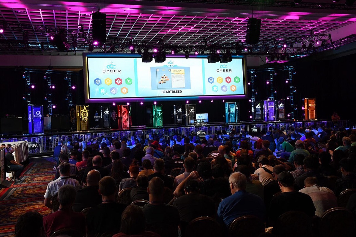 An audience in a dark arena watches large screens showing a live visualization of autonomous hacking systems at DARPA's Cyber Grand Challenge