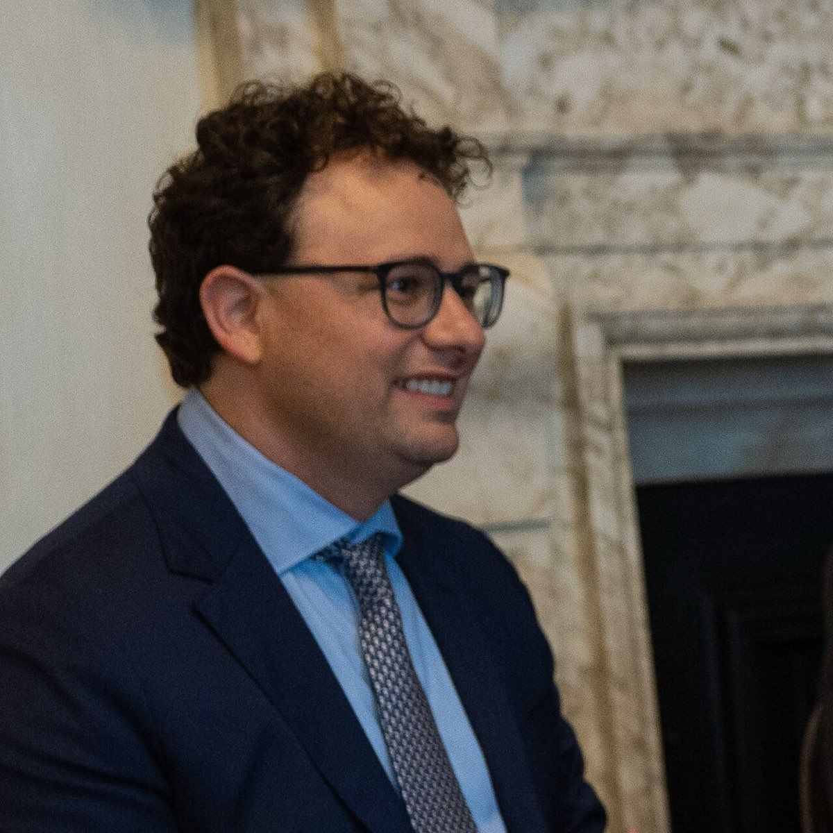 Anthropic chief executive Dario Amodei, wearing glasses and a dark suit, smiling in a room with a marble fireplace