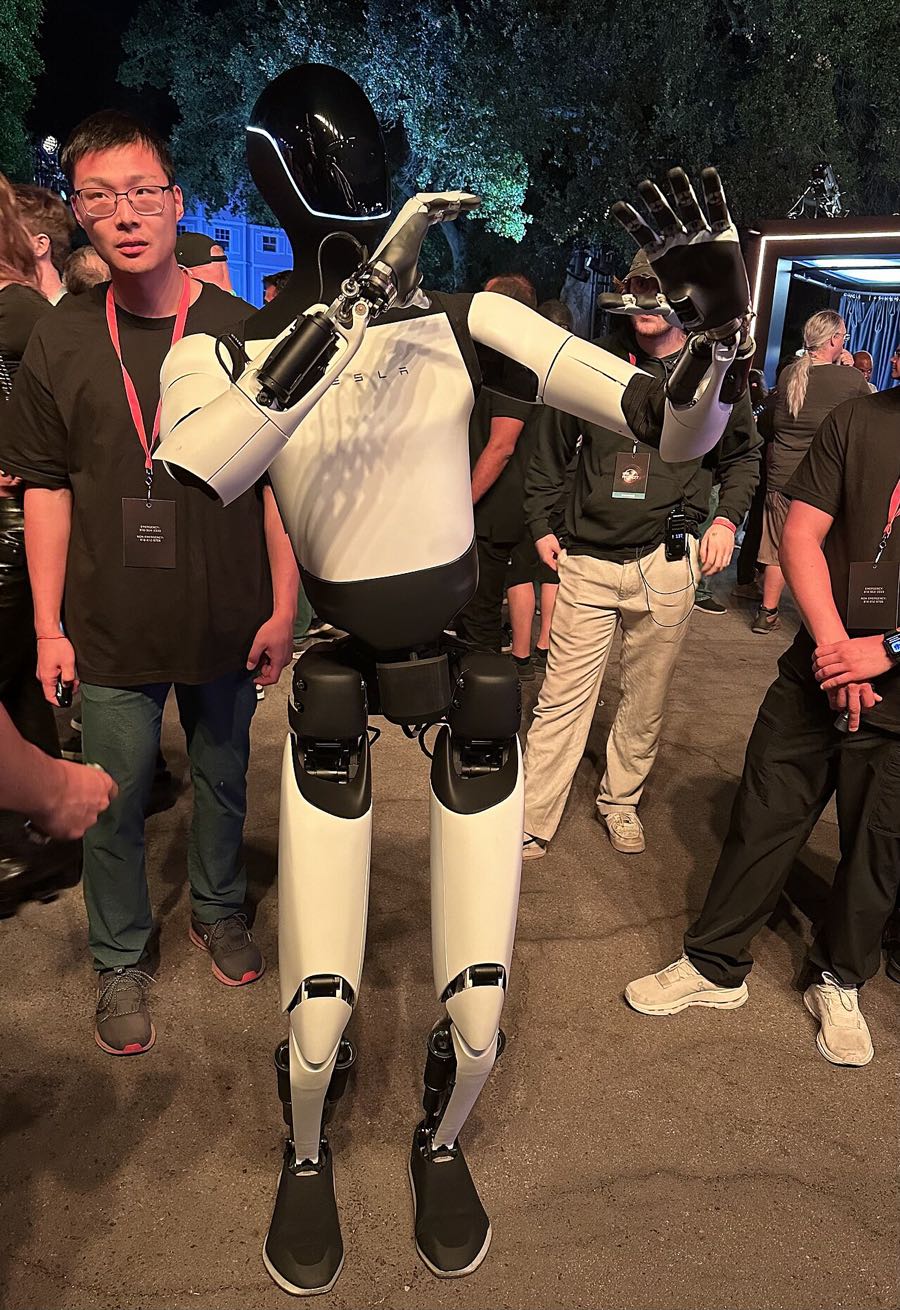 A Tesla Optimus humanoid robot with white limbs and a black head, raising one hand, standing among people at an outdoor evening event