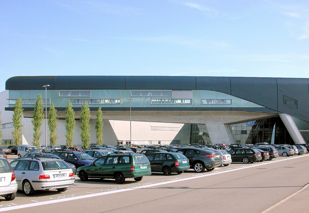 The entrance building of BMW's Leipzig plant: a long, low building with a green metal facade and a sweeping roof, with a parking lot full of cars in front