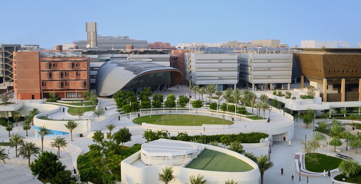 A modern university campus with a curved silver building, terracotta facades and a round central lawn with palm trees