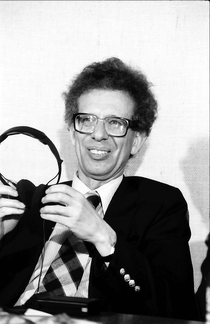 Black-and-white photo of Howard Temin, a smiling man with curly hair and thick glasses in a suit and checked tie, holding a pair of headphones