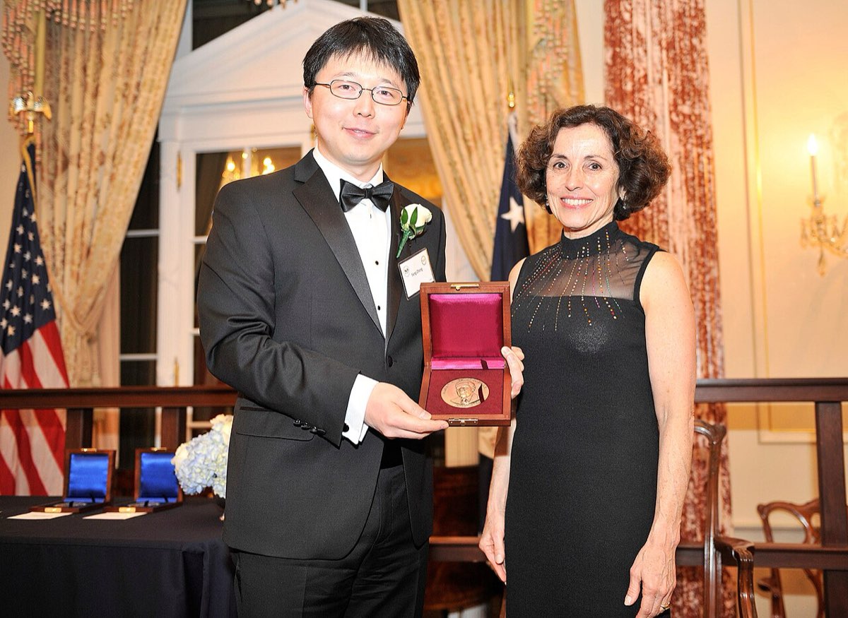 Feng Zhang in a tuxedo holding an open medal case beside NSF Director France Córdova in a formal reception room with flags