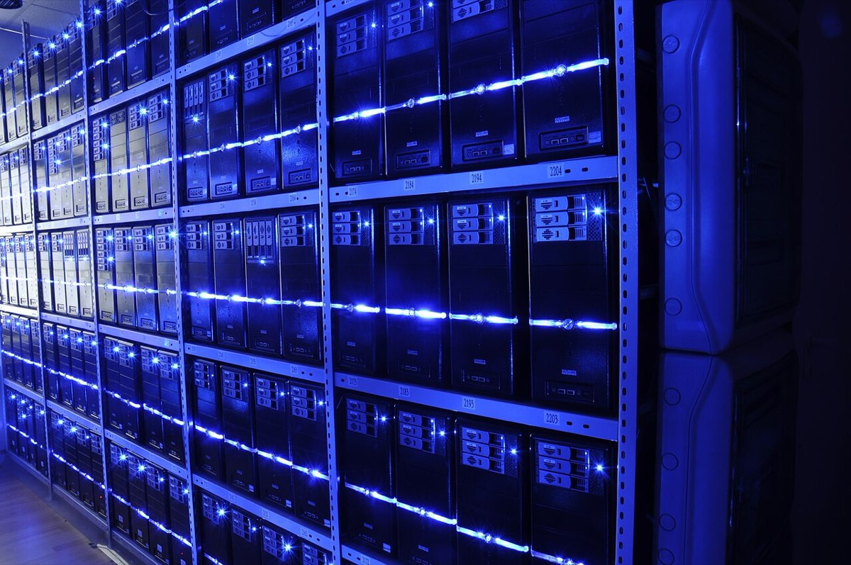 Rows of tower servers on metal shelving in a dark room, lit by blue LED strips
