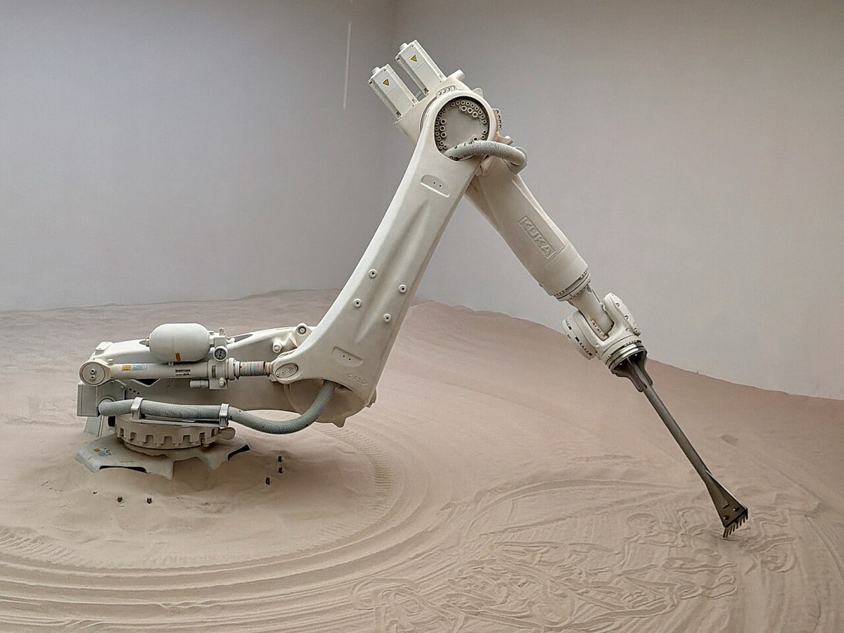 A white industrial robot arm made by KUKA, raking patterns into a floor of sand in a gallery installation
