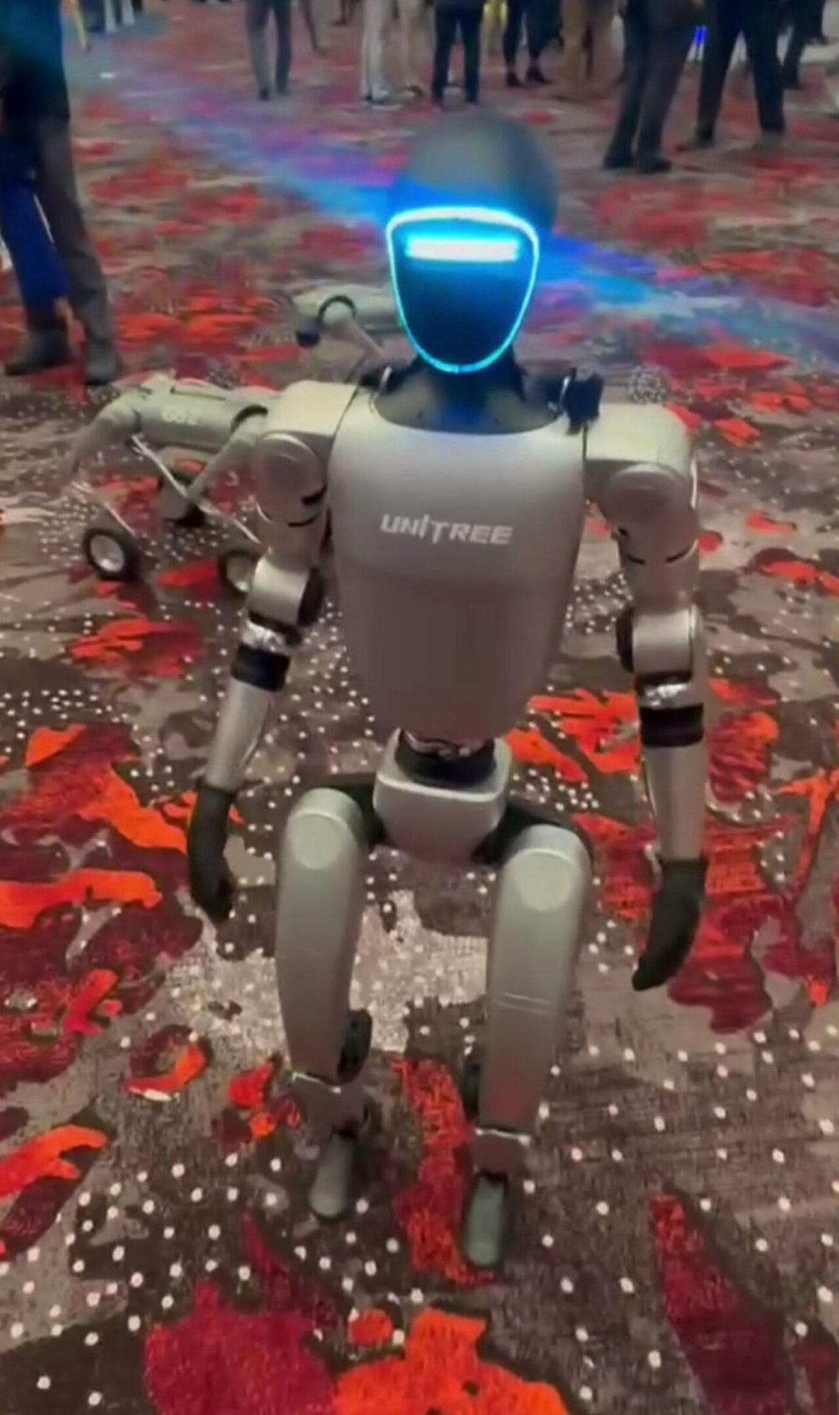 A Unitree G1 humanoid robot with a glowing visor walking on a patterned convention-hall carpet, with a robot dog behind it