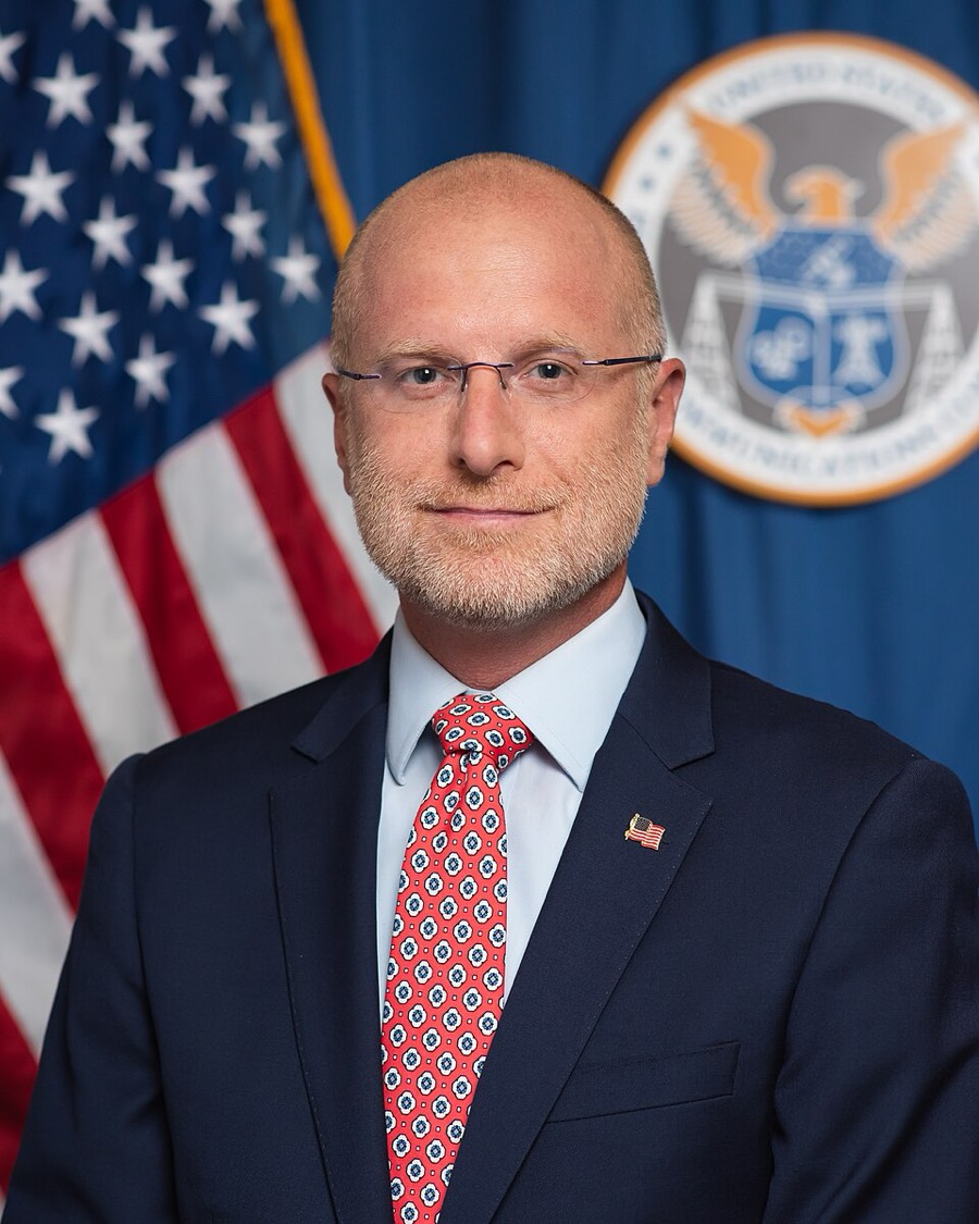 Official portrait of a bald man with glasses and a gray beard, in a dark suit and red patterned tie, in front of an American flag and the FCC seal