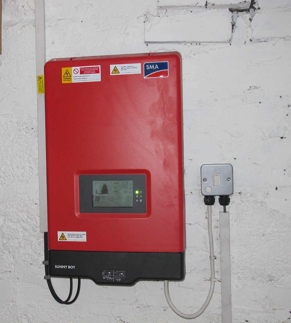 A red wall-mounted solar inverter with a small display, connected by cables to an isolator switch on a whitewashed brick wall