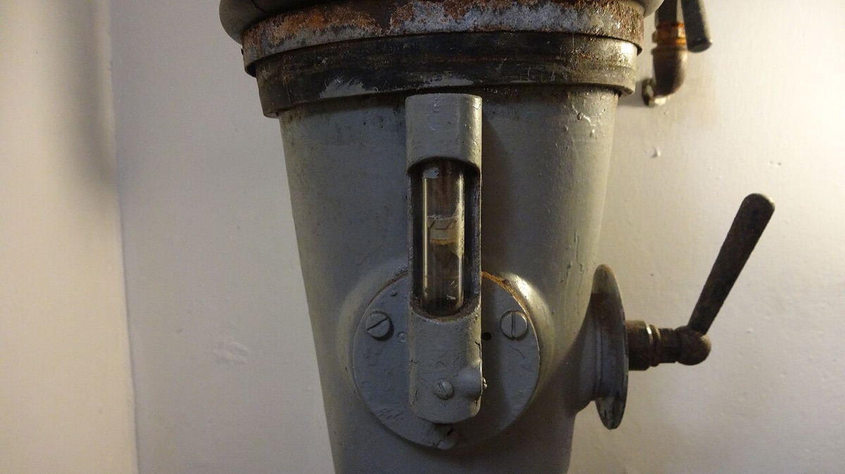 A gray metal duct with a small vertical sight glass and a lever handle, in a Cold War era shelter in Rotterdam