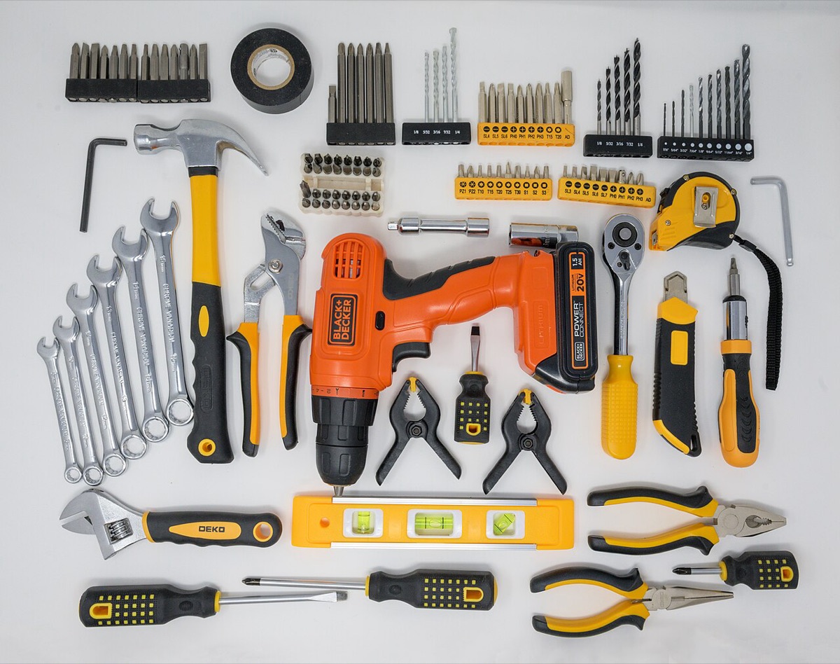 Hand and power tools arranged in rows on a white surface, including a hammer, pliers, wrenches, screwdrivers and a cordless drill