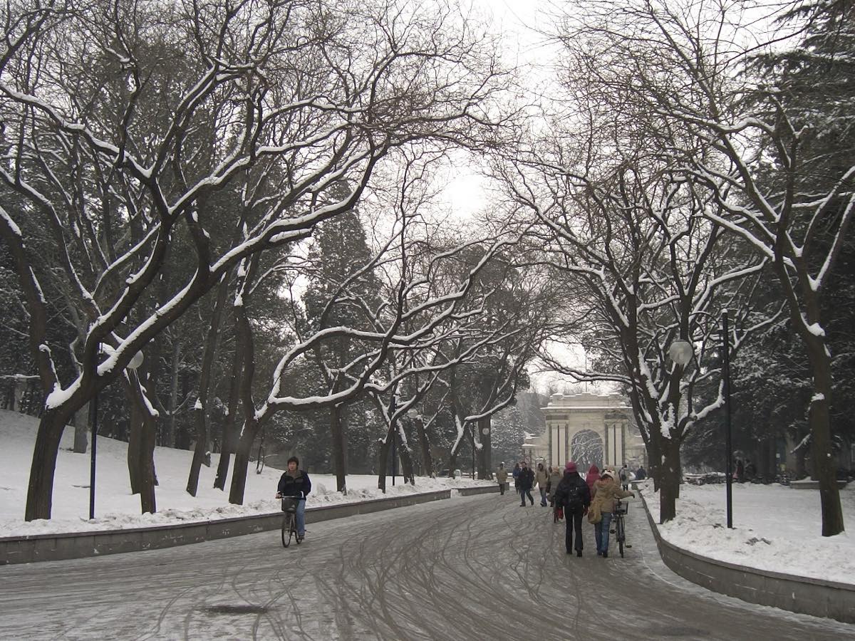 A tree-lined road covered in snow leading to a white stone gate, with people walking and cycling