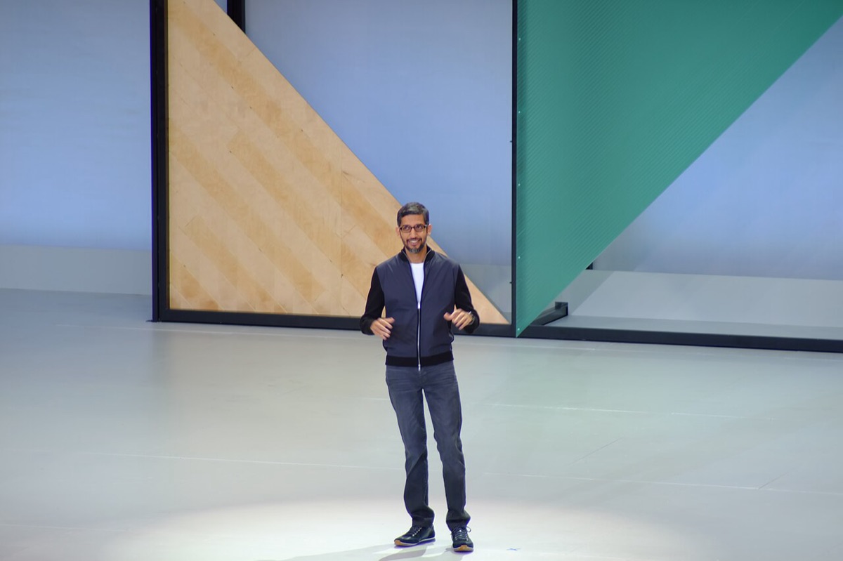 Sundar Pichai standing alone on a large stage with angular wood and green panels behind him