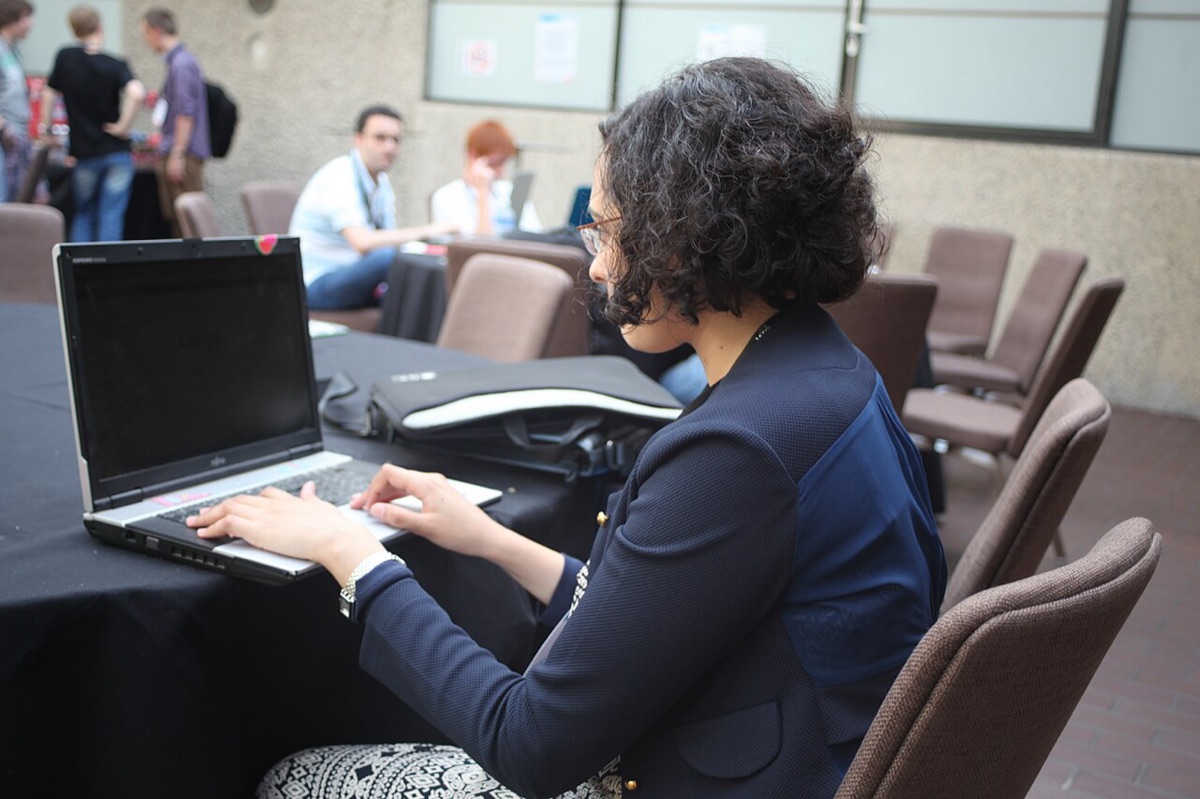 A participant typing on a laptop at a table during a hackathon, with other attendees working in the background