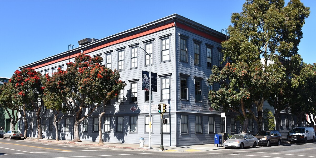 A three-storey grey wooden building with red trim on a sunny street corner, lined with flowering trees