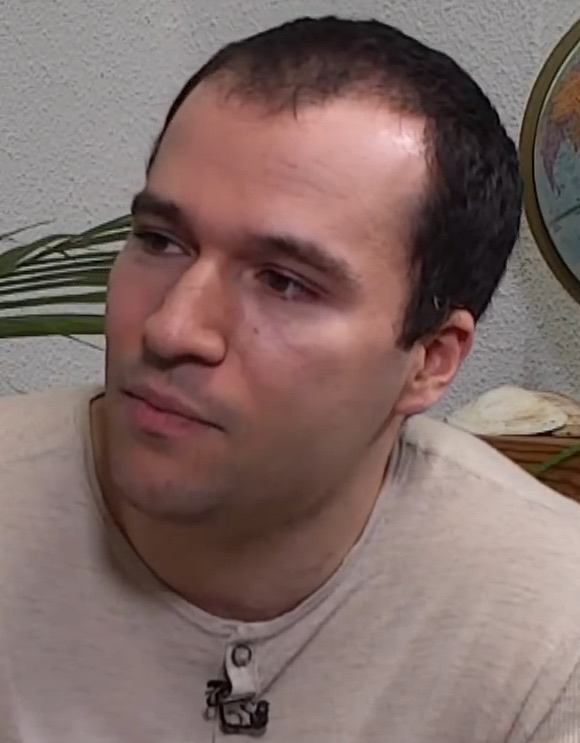 Close-up of Greg Brockman, a man with short dark hair in a light grey shirt, looking to one side during a video interview