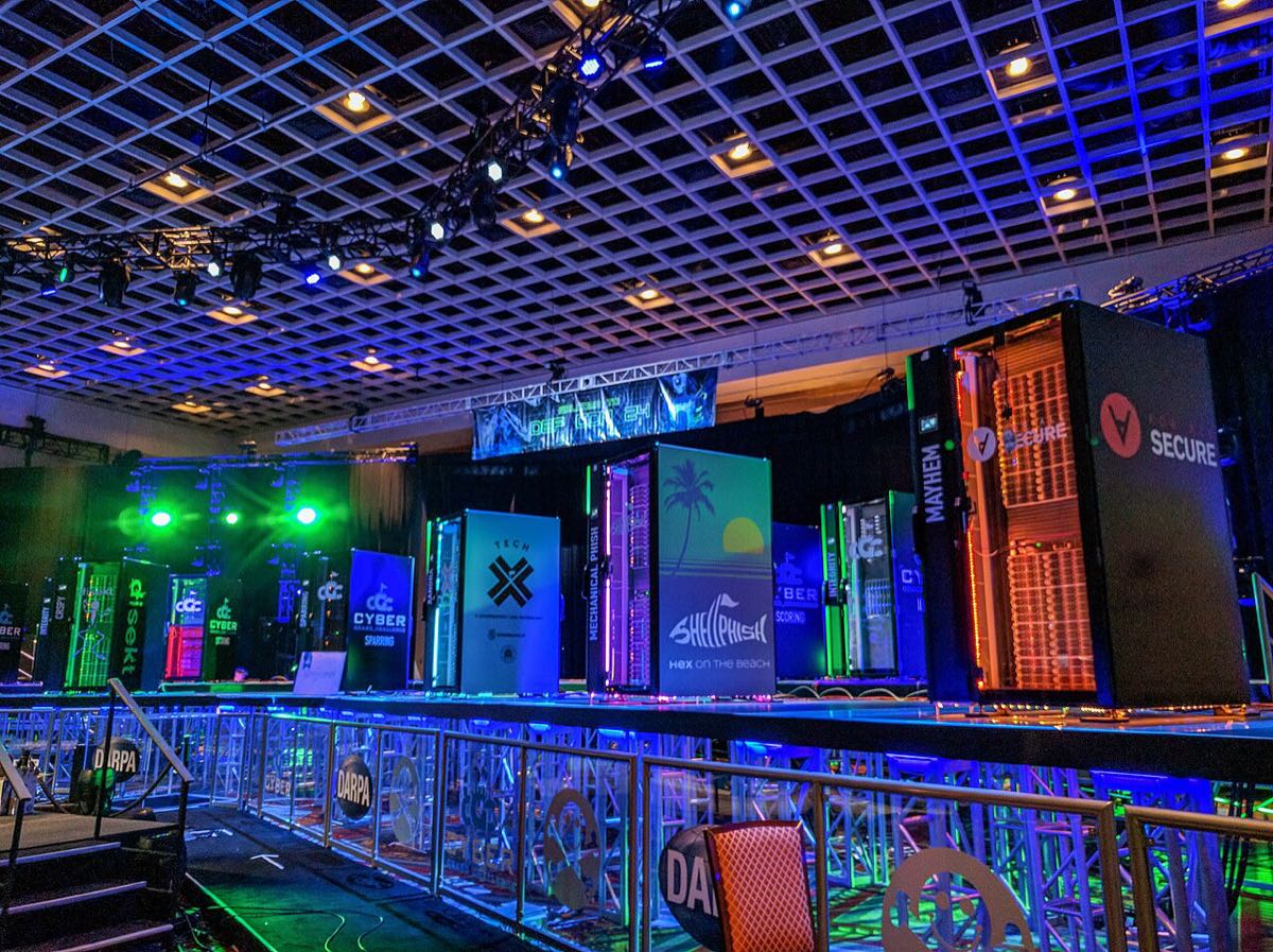Tall server racks with illuminated team branding lined up on a stage under blue and green lights in a conference hall