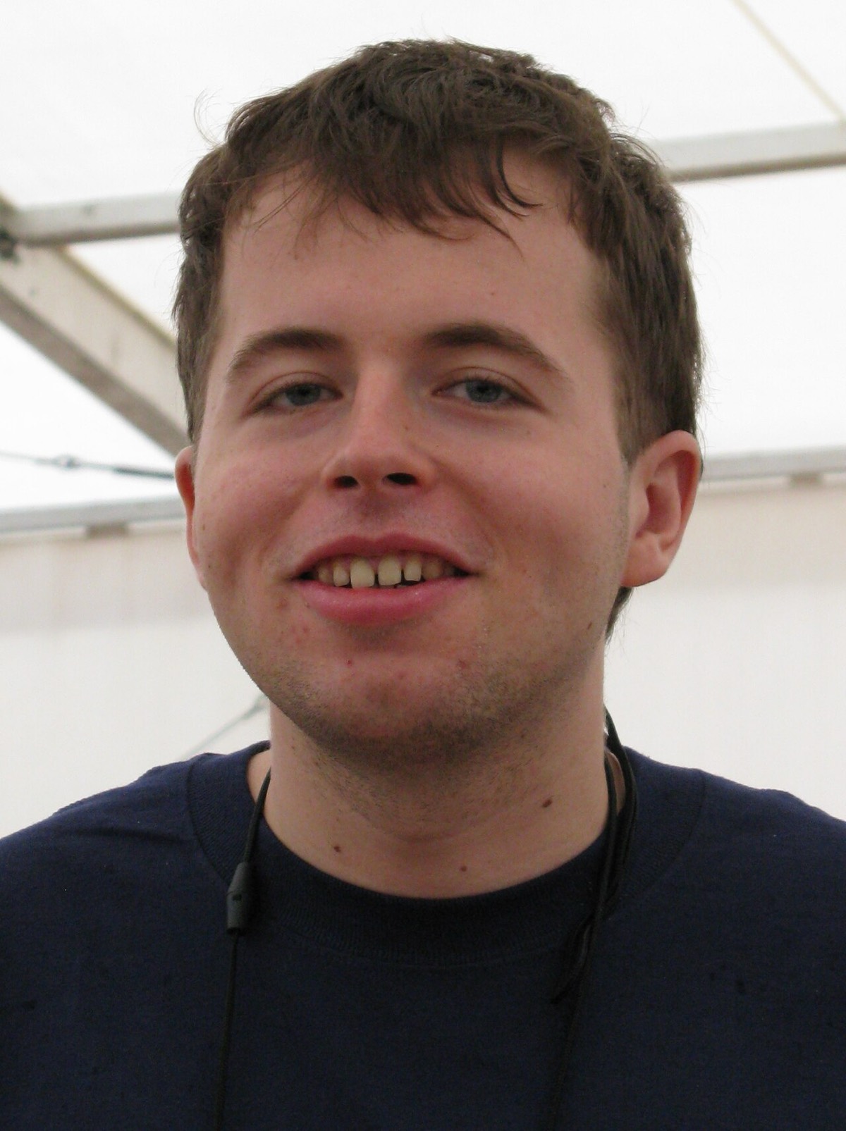 Close-up portrait of a smiling young man with short brown hair in a navy sweater, under a white tent roof