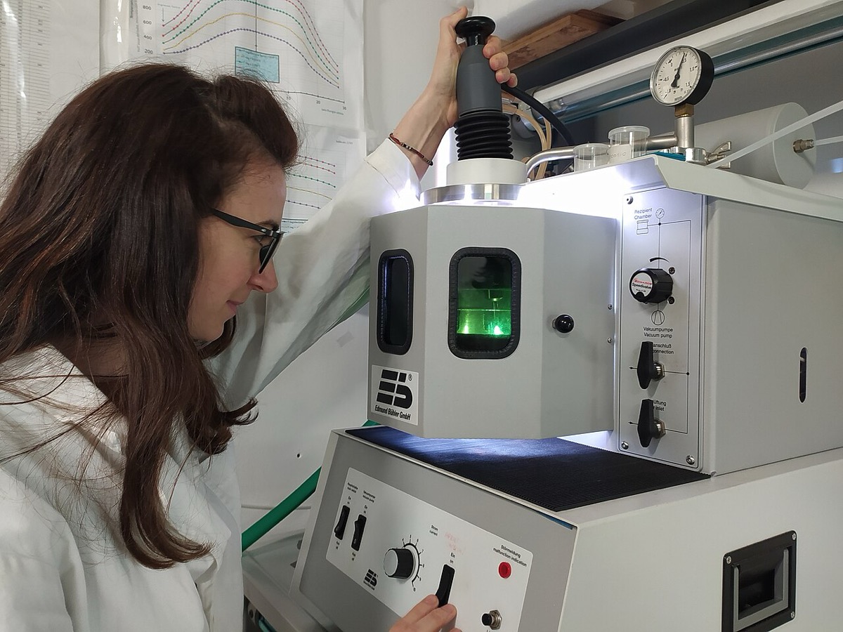 A researcher in a lab coat and glasses operating a compact arc-melting furnace, looking through a small window at a green glow inside the chamber