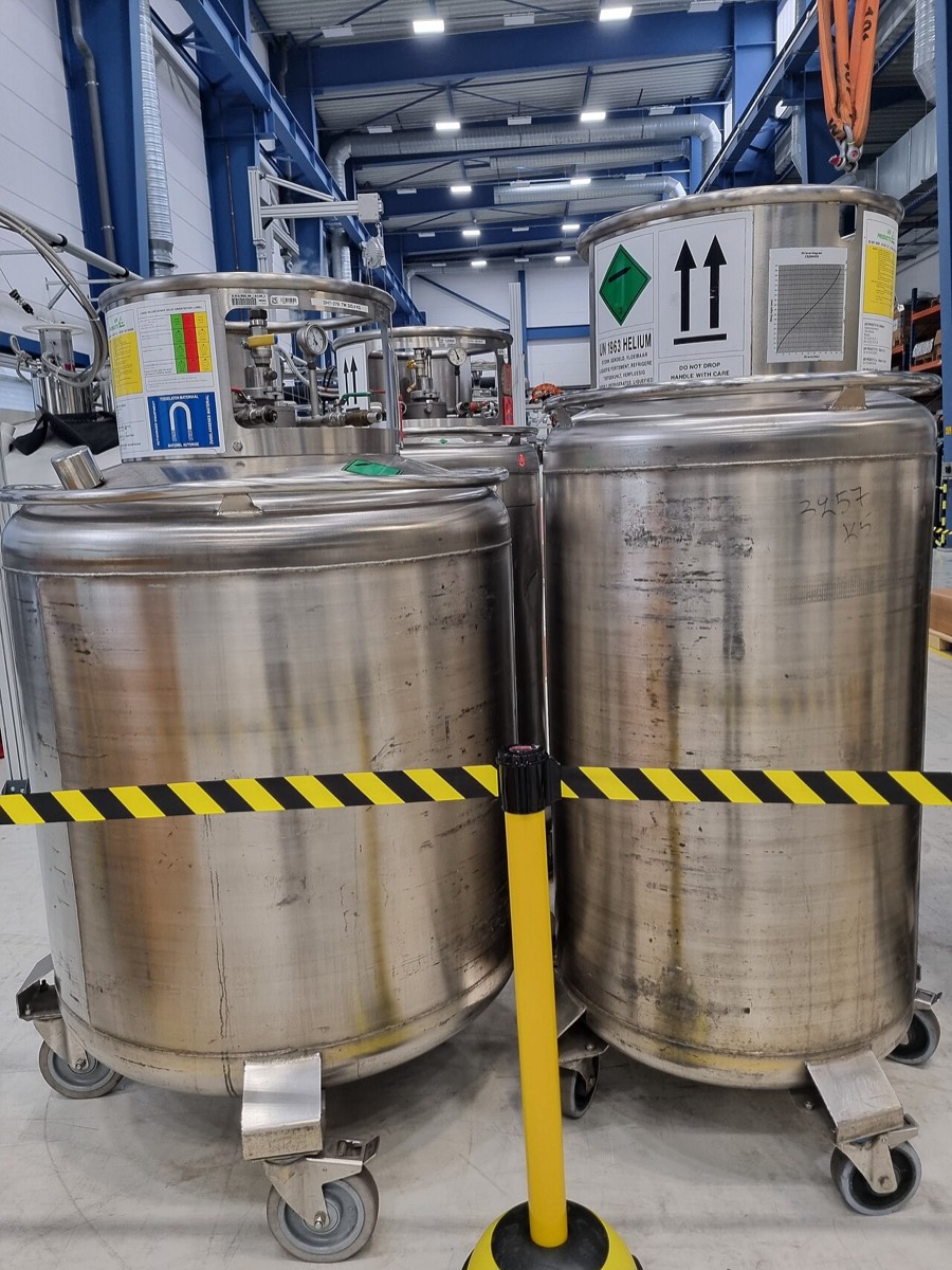 Two large steel liquid-helium dewars on wheels behind a yellow and black barrier tape in a laboratory hall