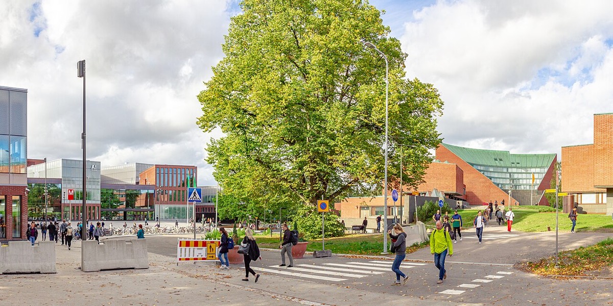 The Otaniemi campus of Aalto University in Espoo: a large tree beside a road crossing, with red-brick and glass university buildings and students walking