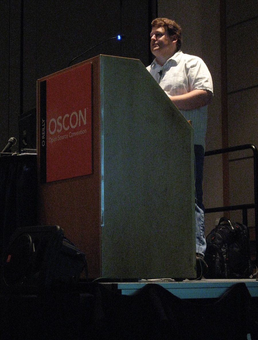 A man in a short-sleeved shirt speaking at a podium marked OSCON on a dark stage