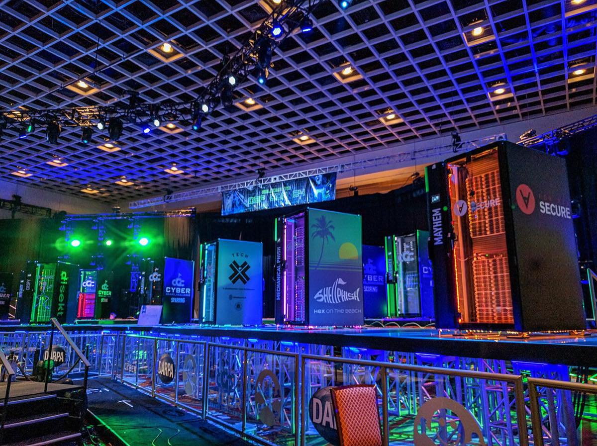 A brightly lit stage at a convention center lined with tall computer racks decorated with team names, behind a railing