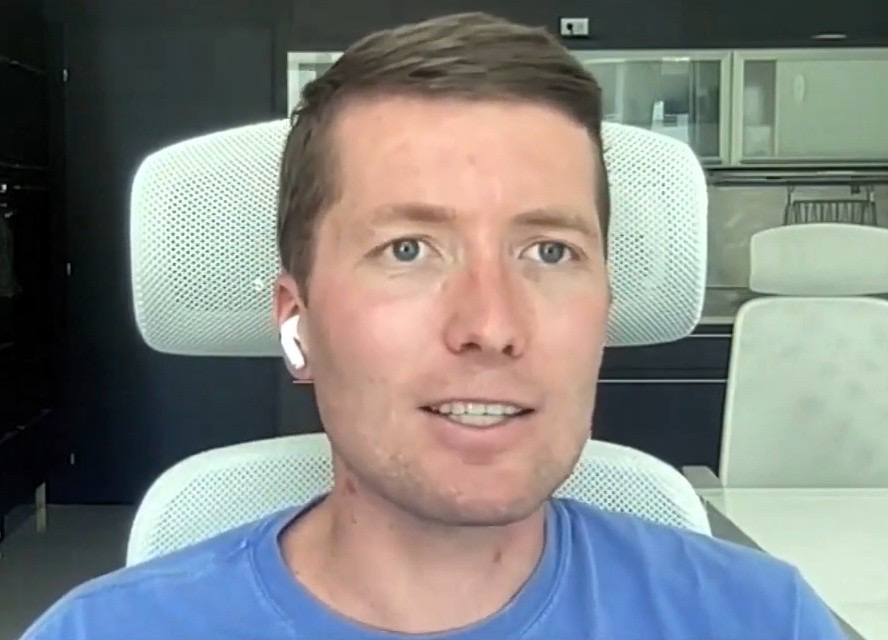 A man in a blue t-shirt speaking on a video call from an office with white chairs