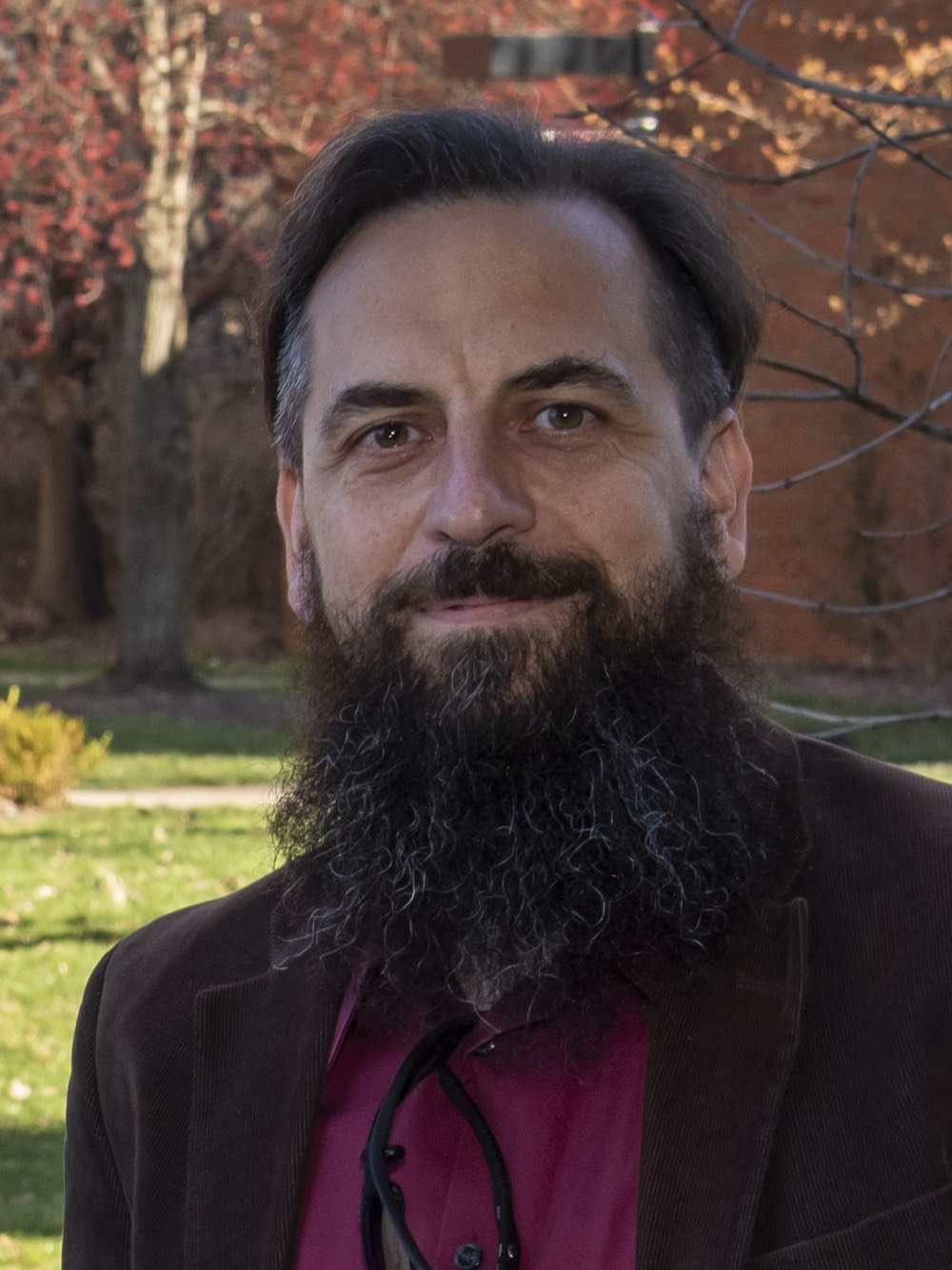 Portrait of a bearded man with dark hair in a dark jacket standing outdoors in front of autumn trees and a brick building