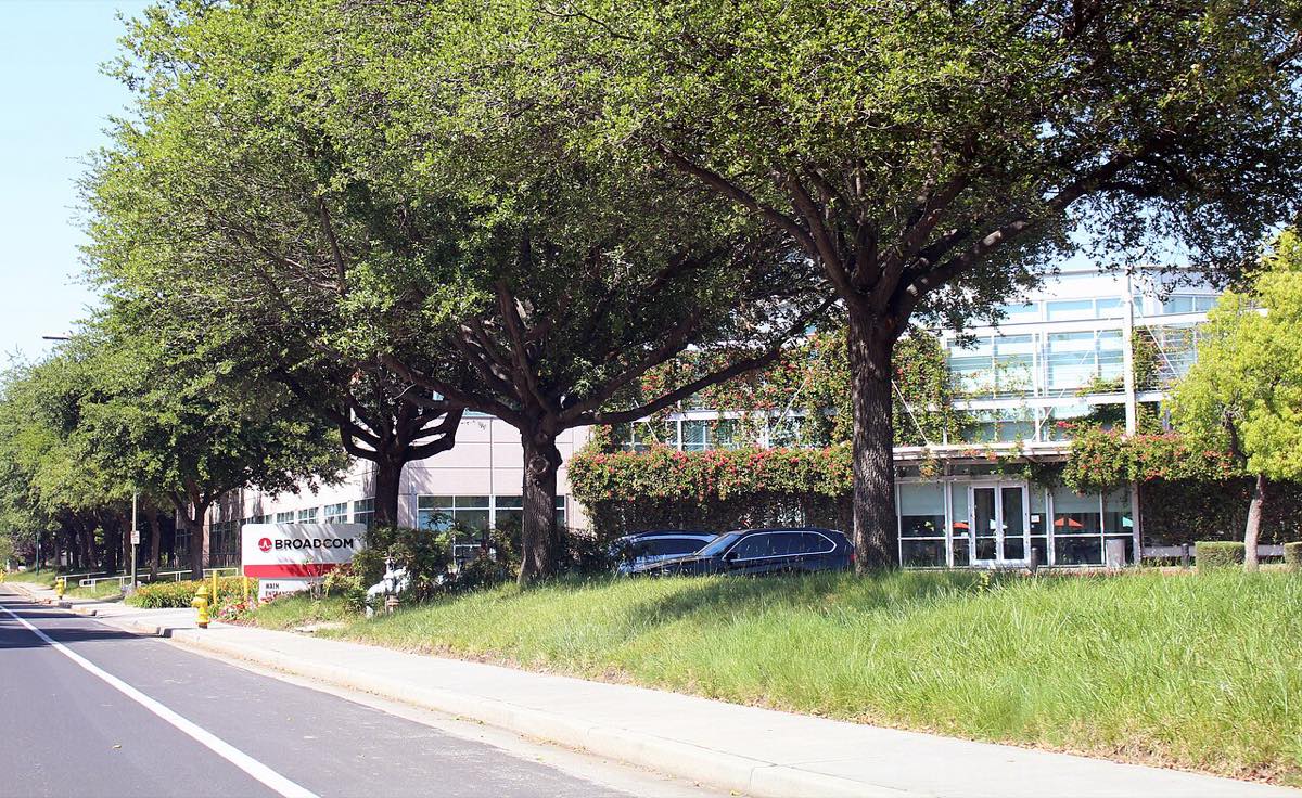 The Broadcom headquarters in San Jose, California: a low office building behind a row of large trees, with a red and white Broadcom sign by the road