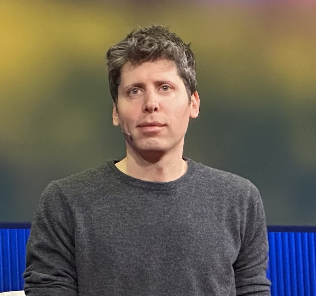 Sam Altman, in a grey sweater, seated on a stage with a blurred yellow and green backdrop