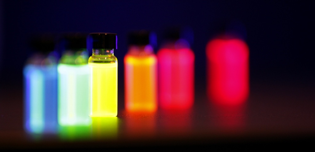 A row of small glass vials glowing blue, green, yellow, orange and red under ultraviolet light