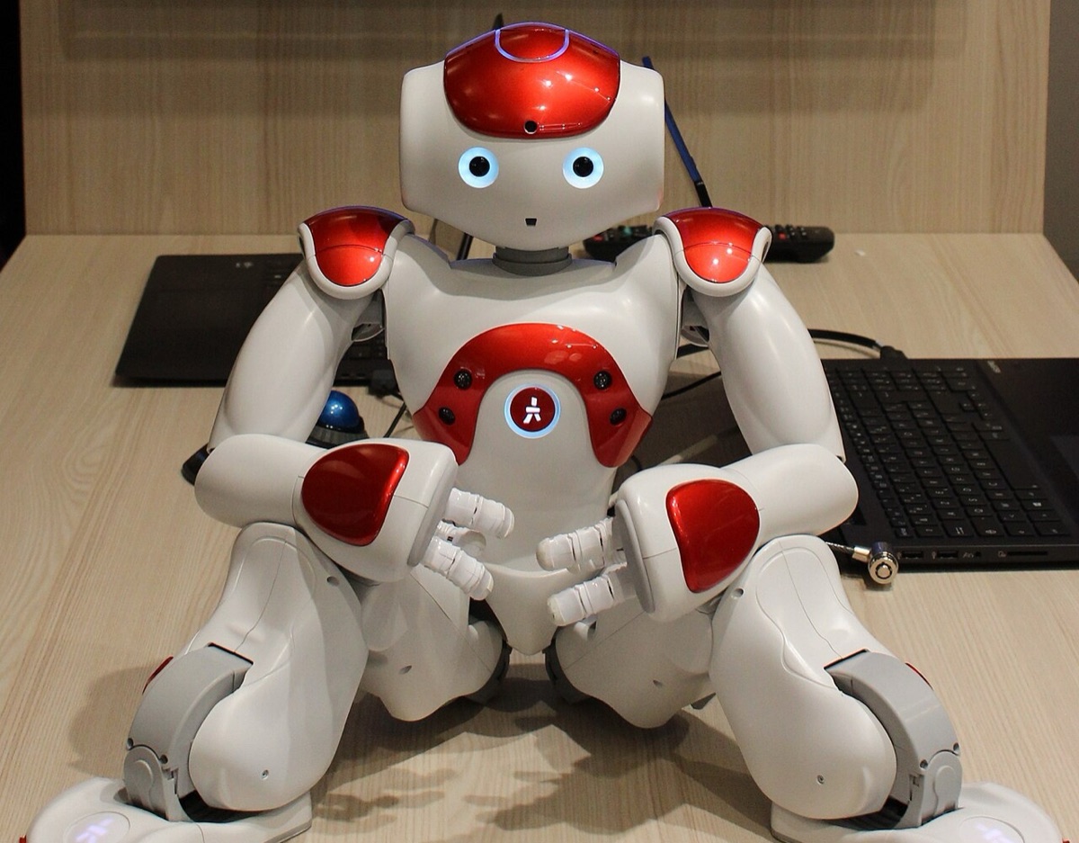 A white and red Nao humanoid robot sitting on a desk in front of two laptops