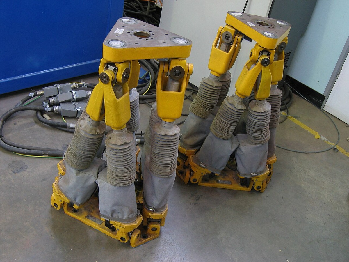 Two yellow industrial hexapod positioners, each a triangular top plate held up by six extendable legs with grey bellows