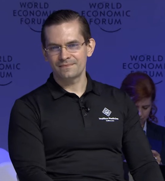 Alex Zhavoronkov, a man with glasses and slicked-back hair in a black polo shirt, seated on a stage in front of a World Economic Forum backdrop