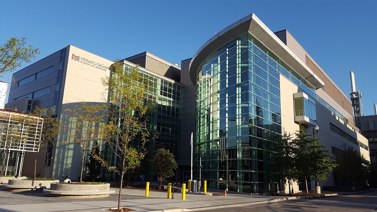 A modern glass and stone medical building at McMaster University on a sunny day, with young trees in front