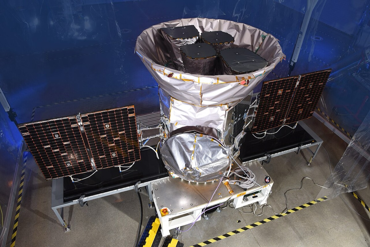 A silver spacecraft with four black camera hoods pointing up inside a conical sunshade, flanked by two dark solar panels, sitting on a test stand in a clean room