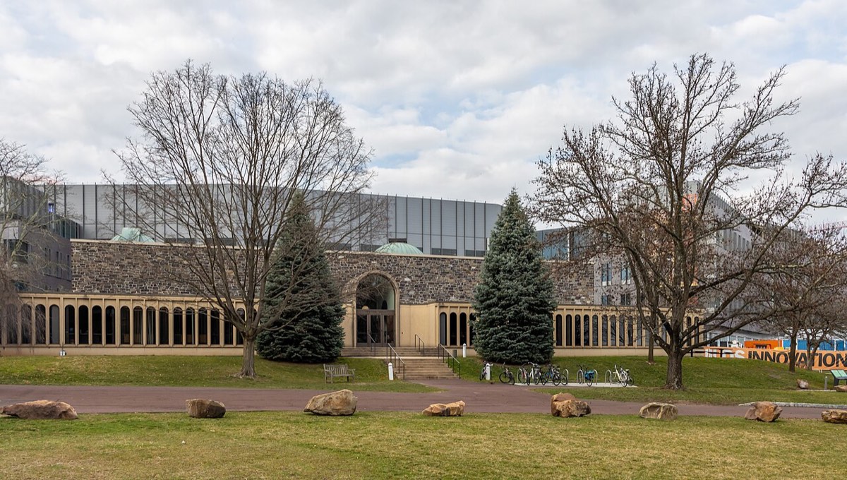 A long, low building with a stone facade and arched entrance behind bare trees and a lawn, with a glass-walled upper floor