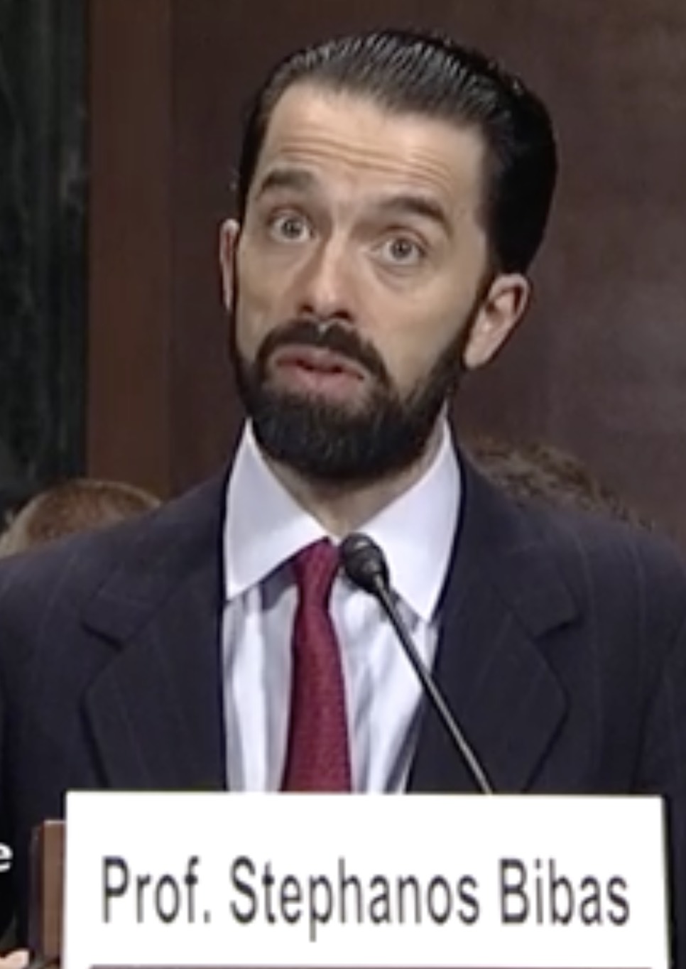 Stephanos Bibas, a man with dark slicked-back hair and a beard, in a dark suit and red tie, speaking at a microphone behind a name placard