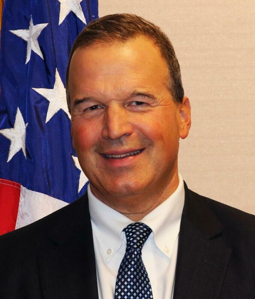 Head-and-shoulders portrait of Judge Luis Felipe Restrepo, a smiling man with short brown hair in a dark suit and blue tie, beside an American flag