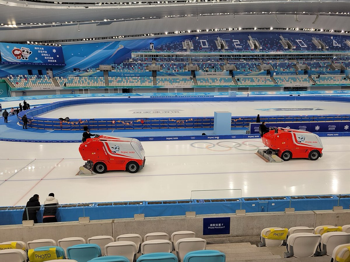 An indoor speed skating rink with two red ice resurfacing machines on the ice and rows of blue seats behind